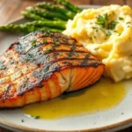 A plated dish of grilled salmon and mashed potatoes, garnished with asparagus and drizzled with butter.